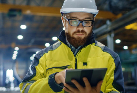 A man in a fluro yellow safety jacket and white hard hat makes notes on a hand held tablet.